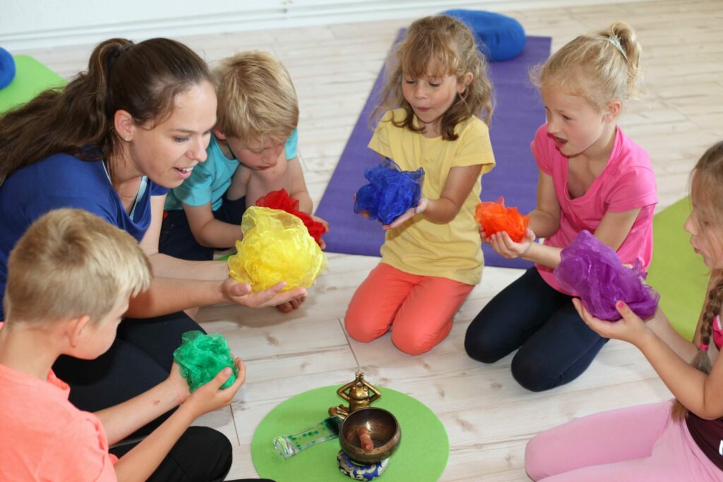 Ulrike sitzt mit vier Kindern im Kreis auf Yogamatten, alle halten bunte Tücher in den Händen